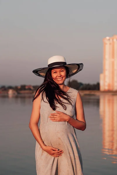 Una mujer embarazada al atardecer. Una mujer embarazada feliz y sonriente con un sombrero de paja y un vestido gris sostiene su vientre embarazada con las manos en un paseo por el río —  Fotos de Stock