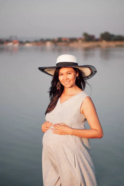 Una mujer embarazada al atardecer. Una mujer embarazada tranquila y relajada con un sombrero de paja y vestido con las manos sobre su estómago embarazada al atardecer junto al mar. —  Fotos de Stock
