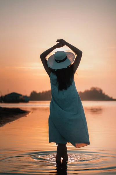 日落时一个女人的轮廓。夏日黄昏时分，一位戴草帽、双手高高地高举在海滩上的年轻女子的背影 — 图库照片