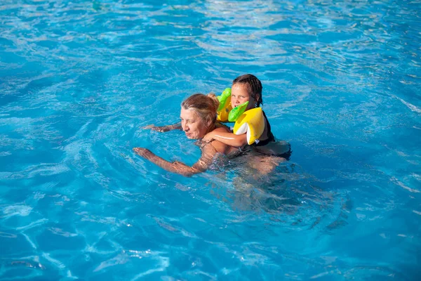 Grandmother and granddaughter in the pool. A girl in inflatable sleeves rides on the back of a young grandmother on a sea family vacation