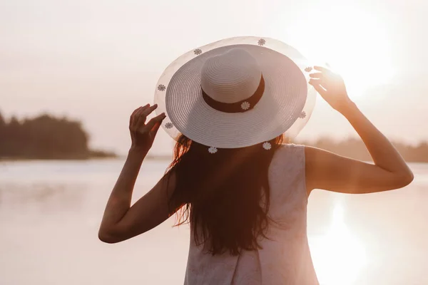 一个夏日的早晨，一位年轻女子手握草帽帽檐，在森林的湖面上欣赏日出，她的背影映入眼帘 — 图库照片