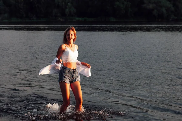 Zdjęcie młodej kobiety w wodzie o zachodzie słońca. Młoda piękna blondynka w białej bluzce i dżinsowych spodenkach biegnie po wodzie na tle pięknego krajobrazu w promieniach słońca — Zdjęcie stockowe