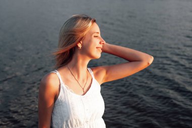 Suyun arka planında profili olan genç bir kadın. Beyaz elbiseli güzel bir sarışının portresi ve gün batımında nehrin kenarında altın mücevherler.