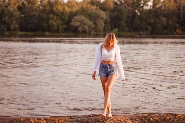 Foto de la mujer en la playa por el río. Una joven rubia con una blusa blanca y pantalones cortos de mezclilla y camisa se para en la arena sobre el fondo de un río y un hermoso paisaje y mira hacia abajo a la arena —  Fotos de Stock