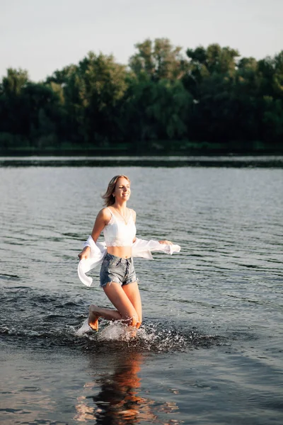 Piękna kobieta biegnie swobodnie przez wodę. Uśmiechnięta młoda blondynka w białej bluzce i dżinsowych spodenkach na tle krajobrazu — Zdjęcie stockowe