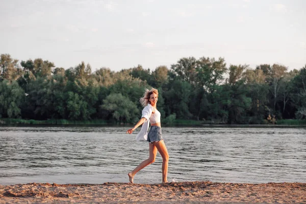 Una joven corriendo contra el fondo del río. Una hermosa rubia feliz en una camiseta blanca y pantalones cortos de mezclilla corre por la playa —  Fotos de Stock