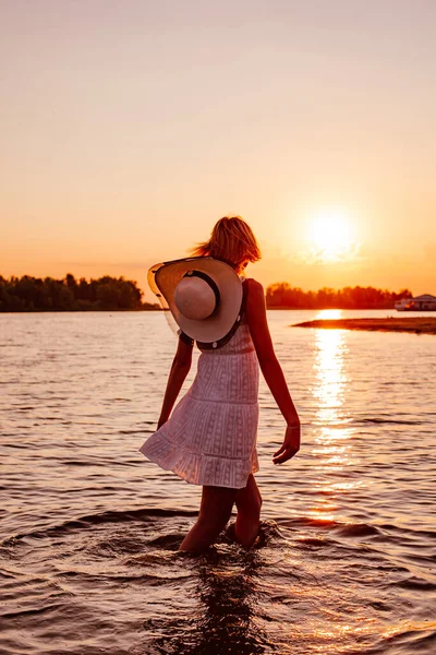 Uma jovem no fundo do pôr-do-sol. Foto visão traseira de uma bela loira feliz em um vestido branco e um chapéu de palha com uma fita escura andando na água e olhando para o pôr do sol — Fotografia de Stock