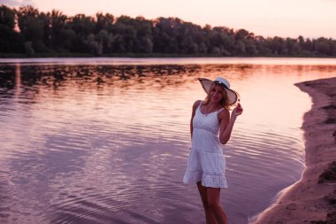 Hasır şapkalı bir kadın poz veriyor. Beyaz dantel elbiseli, genç, güzel bir sarışın gün batımına karşı nehir kıyısında duruyor ve şapkasını tutuyor.