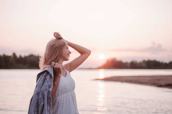 Retrato de perfil de una joven en la playa. Una hermosa rubia en un vestido blanco de verano se encuentra en la orilla del río y sostiene una chaqueta de mezclilla en su mano contra el fondo de la puesta del sol y el agua —  Fotos de Stock