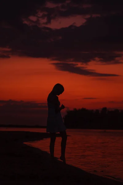 Sylwetka kobiety z profilem na tle zachodu słońca. Smukła młoda kobieta spaceruje wzdłuż brzegu rzeki na tle chmur i plaży — Zdjęcie stockowe