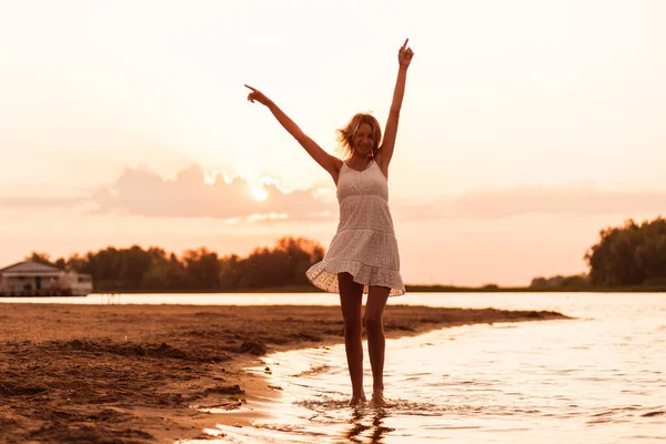 Uma jovem está dançando contra o fundo do pôr do sol. Uma bela loira delgada feliz em um vestido de verão branco está girando na areia contra o pano de fundo de uma paisagem pitoresca — Fotografia de Stock