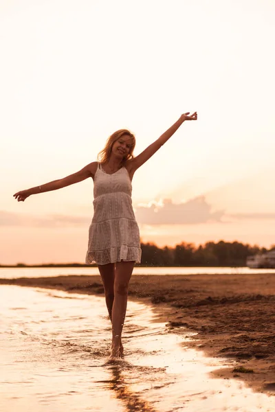 Hermosa mujer posando en la playa. Una joven rubia delgada con un vestido blanco está girando en la orilla del río contra el fondo del atardecer y levanta las manos —  Fotos de Stock