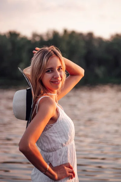 Retrato de una rubia sobre el fondo de un río. Una hermosa joven feliz posa al atardecer con un vestido blanco y un sombrero de paja con una cinta oscura sobre el fondo de los árboles —  Fotos de Stock