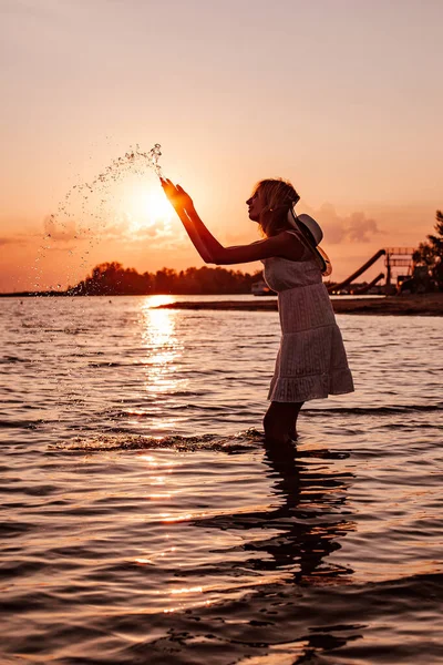 Uma jovem espirra água. Foto de uma loira delgada, bonita e feliz em um vestido de verão e um chapéu de palha em pé no rio contra o fundo da praia e do pôr do sol — Fotografia de Stock