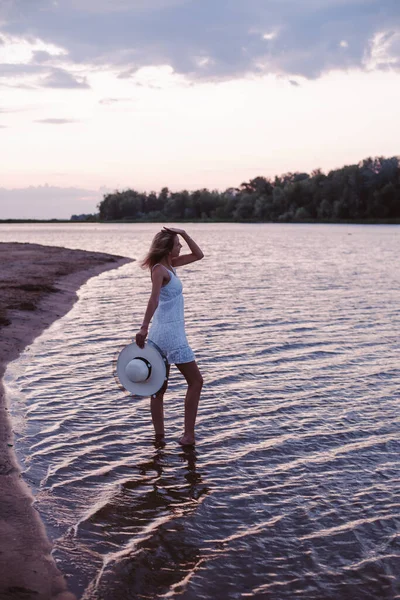 Uma jovem olha para o pôr-do-sol. Uma bela loira delgada feliz está de pé na margem do rio em um vestido branco e segurando um chapéu de palha em sua mão — Fotografia de Stock