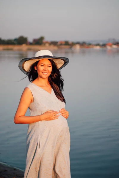 Una mujer embarazada al atardecer. Hermosa mujer embarazada asiática en un sombrero de paja con las manos en su vientre embarazada disfruta de la puesta de sol junto al río —  Fotos de Stock