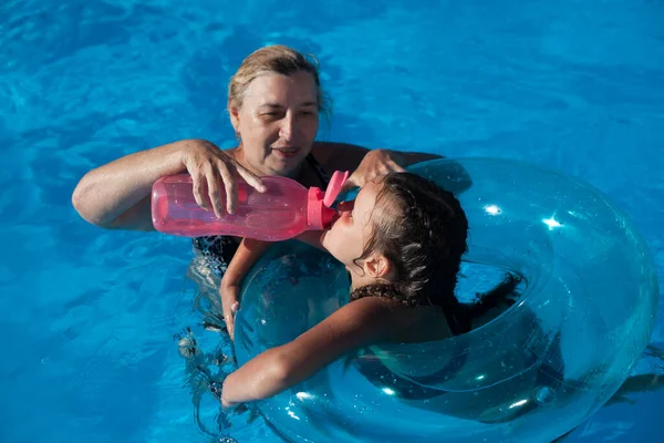 Grandmother and granddaughter in the pool. A young grandmother gives her granddaughter water from a reusable plastic drinking bottle in the swimming pool on summer vacation