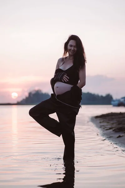 Беременная женщина в черном костюме. Красивая сексуальная молодая женщина в шелковой пижаме стоит на одной ноге в воде на берегу моря на фоне заходящего солнца — стоковое фото