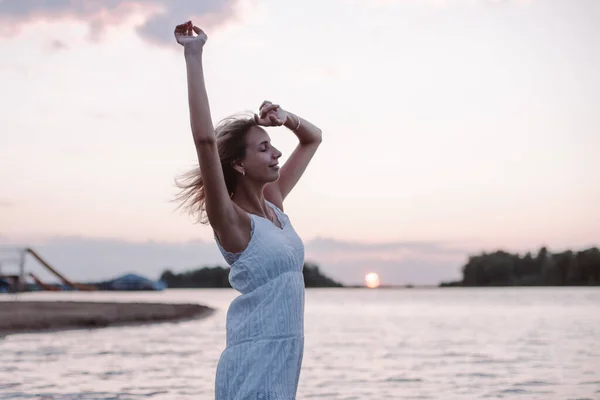 Una joven posa sobre el fondo de un paisaje. Foto de una hermosa rubia feliz en un vestido de verano blanco con su pelo soplando en el viento en la orilla del río y sus manos levantadas —  Fotos de Stock