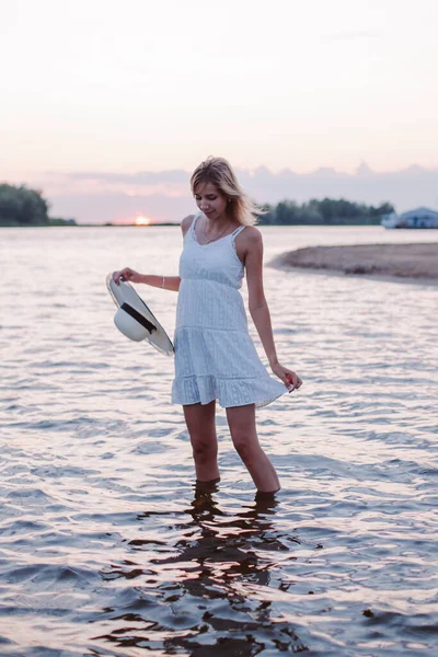 Młoda kobieta stoi w wodzie. Piękna szczęśliwa blondynka w białej letniej sukience na tle zachodu słońca trzyma słomkowy kapelusz i rąbek sukienki w rękach — Zdjęcie stockowe
