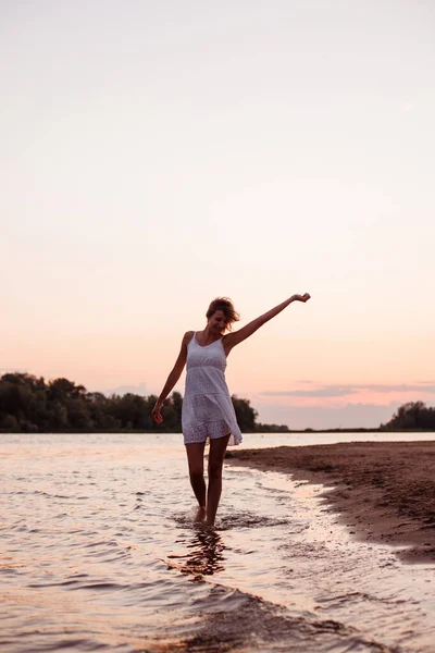Foto de uma dançarina ao pôr-do-sol. Loira bonita em um vestido de verão branco na margem do rio contra o fundo da praia e da praia — Fotografia de Stock