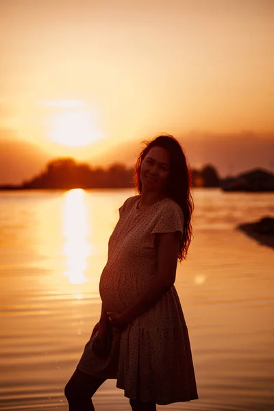 Mujer joven al atardecer. Silueta de mujer feliz embarazada con el pelo largo en rayos de sol poniente, de pie en la playa y disfrutando del paisaje de verano —  Fotos de Stock