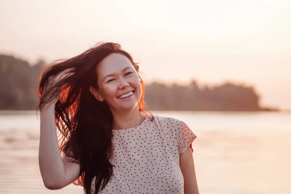 Mujer sonriente al aire libre. Retrato de cerca de la mujer asiática feliz sobre el fondo de la playa y el mar, jugando con el pelo largo y oscuro y disfrutando de la puesta de sol —  Fotos de Stock