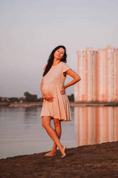 Gravid brunette på baggrund af byen. Smuk ung asiatisk kvinde med langt brunt hår står barfodet på stranden i sommerkjole nær højhuse - Stock-foto