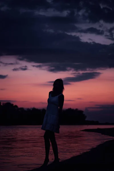 Γυναίκες ποζάρουν στο φόντο του νερού και του ηλιοβασιλέματος. Μια λεπτή νεαρή γυναίκα στέκεται σε ένα καλοκαιρινό φόρεμα στην όχθη του ποταμού με φόντο τα σύννεφα και το τοπίο — Φωτογραφία Αρχείου