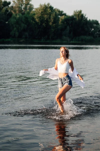 Piękna młoda kobieta biegnie swobodnie przez wodę. Młoda blondynka w białej bluzce i dżinsowych spodenkach na tle drzew i rzeki — Zdjęcie stockowe