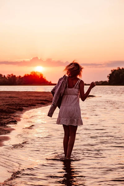 Uma jovem está girando contra o fundo do pôr-do-sol. Uma bela jovem loira em um vestido de verão branco e uma jaqueta de ganga em sua mão está de pé na água na margem do rio — Fotografia de Stock