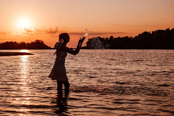 Μια νεαρή γυναίκα στο φόντο του ηλιοβασιλέματος. Φωτογραφία από μια σιλουέτα μιας όμορφης ξανθιάς με λευκό καλοκαιρινό φόρεμα και ψάθινο καπέλο που πιτσιλάει νερό και ποζάρει στην παραλία με θέα το ηλιοβασίλεμα — Φωτογραφία Αρχείου