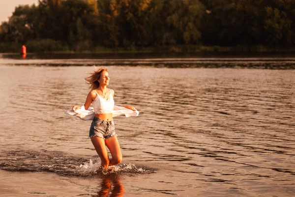 Eine junge Frau läuft vor dem Hintergrund des Flusses und der Morgendämmerung. Eine lächelnde Blondine in weißem Top, Hemd, Jeanshose rennt im Wasser vor einer wunderschönen Landschaft — Stockfoto