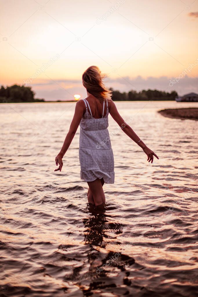 A young woman is standing in the water from behind. A beautiful blonde in a white summer dress poses against the sunset and raises her arms to the sides