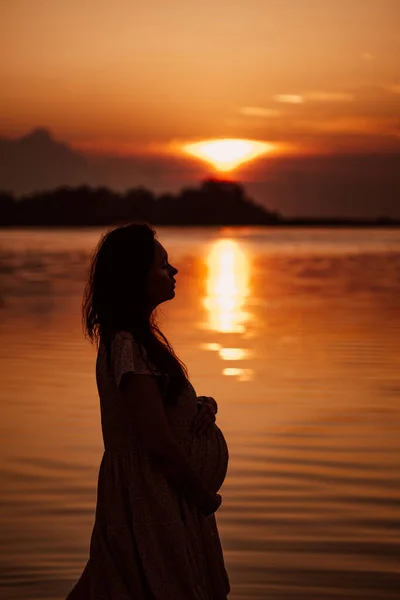 在日落的时候，怀孕的女人。站在海滨的美丽女子的水中反射出夕阳西下的侧影 — 图库照片