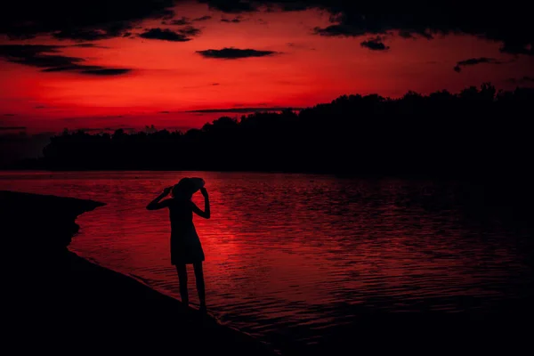 夕阳西下,一个女人在海滩背景下的轮廓.一位纤细的年轻女子站在河岸上，手里拿着一顶帽子 — 图库照片