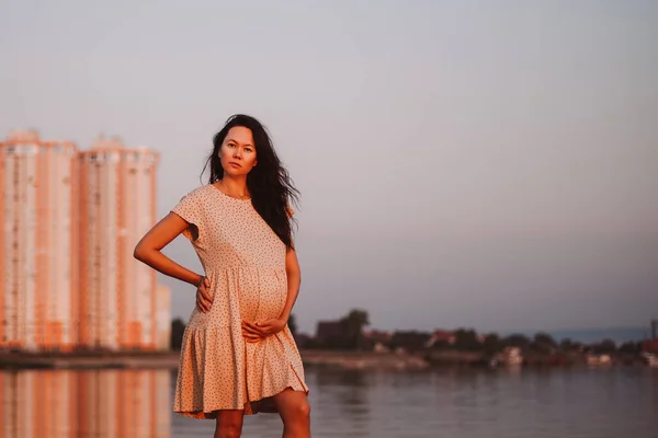 La morena embarazada posa junto al lago. Joven mujer asiática sostiene su vientre embarazada con sus manos y disfruta de los rayos de sol poniente sobre el fondo de edificios de gran altura —  Fotos de Stock