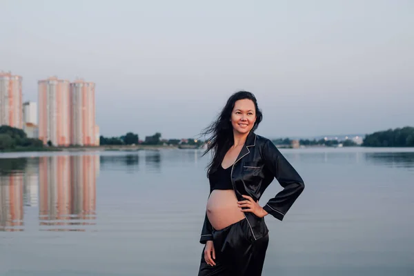 Улыбающаяся беременная брюнетка у воды. Молодая красивая сексуальная женщина в темном шелковом костюме позирует на фоне высотных зданий с рукой на талии — стоковое фото