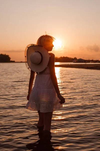 Uma jovem no fundo do pôr-do-sol. Uma foto de perfil de uma linda loira em um vestido branco e um chapéu de palha com uma fita em torno de seu pescoço em pé na água — Fotografia de Stock