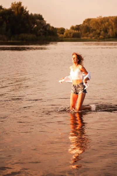 Młoda kobieta biegnie do wody o świcie. Piękna uśmiechnięta blondynka w białej bluzce i opuszczonej koszuli, jeansowe szorty biegną swobodnie w rzece na tle krajobrazu — Zdjęcie stockowe