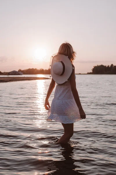 日落时一个年轻女子的照片。一位身材苗条、美丽的金发姑娘，身穿白色的夏装，头戴草帽，站在水里，凝视着夕阳和美丽的风景 — 图库照片