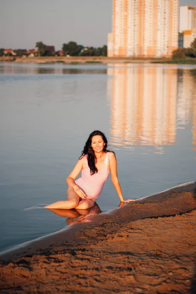 Mujer embarazada en la playa, estilo de vida de larga duración retrato de una mujer embarazada en un traje de baño rosa posando en una playa de río de arena con edificios de varios pisos en el fondo —  Fotos de Stock