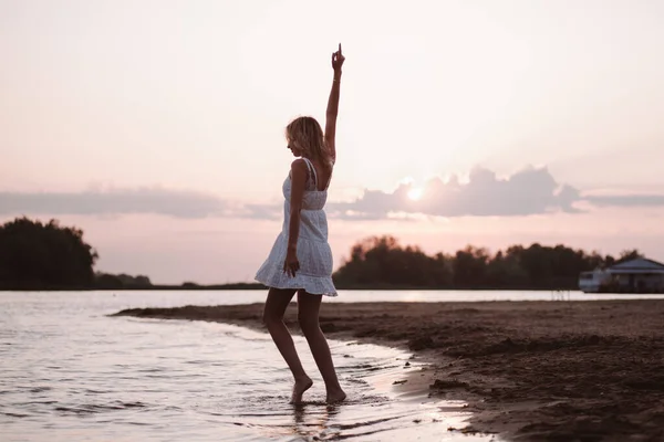 Una joven está bailando en la playa. Una hermosa rubia feliz delgada en un vestido de encaje blanco está girando en el agua contra el fondo de una puesta de sol y un paisaje pintoresco —  Fotos de Stock