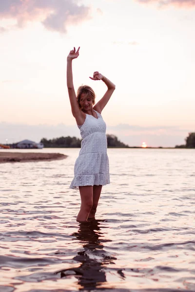 Una joven está bailando en el agua. Foto de una hermosa rubia feliz en un vestido de verano blanco y con las manos levantadas bailando sobre el fondo del atardecer —  Fotos de Stock