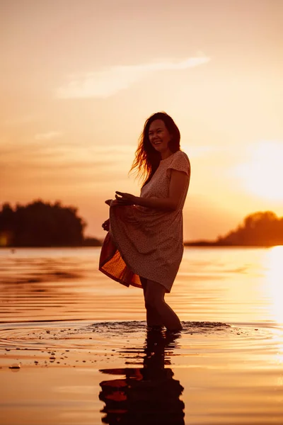 Silhouette einer Frau bei Sonnenuntergang. Junge glückliche Frau steht am Sommerabend im Meer in den Strahlen der untergehenden Sonne, hält den Saum ihres Kleides in den Händen und lächelt — Stockfoto