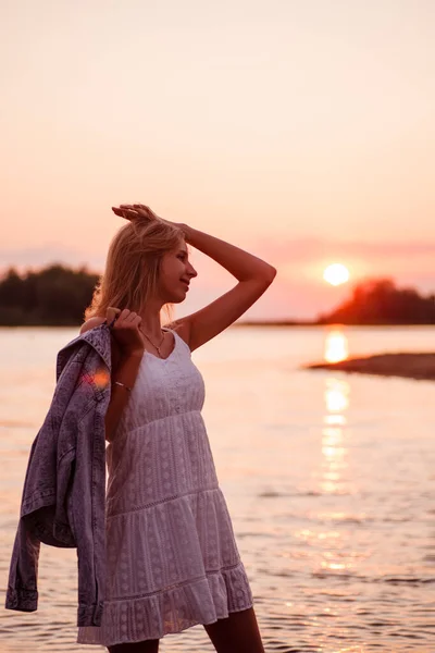 Foto en el perfil de una joven. Una hermosa rubia feliz en vestido de verano blanco se para en la orilla del río y sostiene una chaqueta de mezclilla en su mano contra el fondo de la puesta de sol y el bosque —  Fotos de Stock