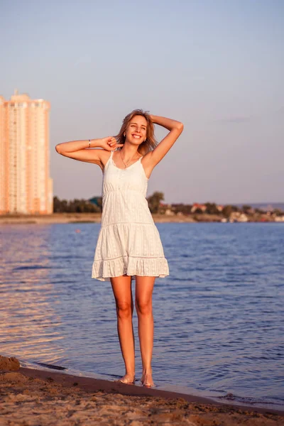 Una joven con un vestido en el fondo de la ciudad. Foto de una hermosa rubia bronceada en un vestido de encaje blanco sonriendo y de pie sobre la arena cerca del río —  Fotos de Stock