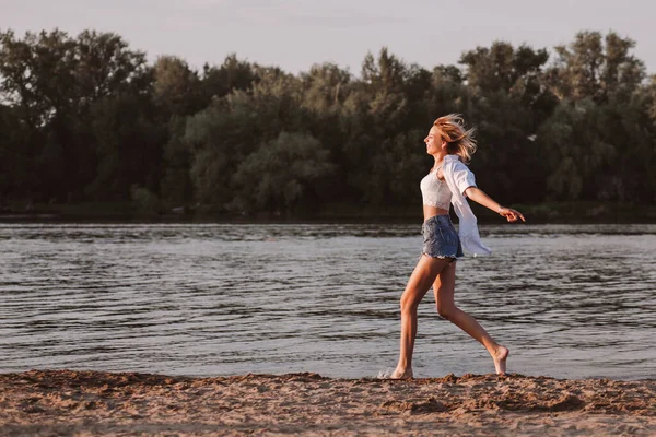 Una joven mujer corriendo en libertad contra el fondo del río y los árboles. Foto de perfil una hermosa rubia feliz en ropa blanca y pantalones cortos de mezclilla corre sobre la arena —  Fotos de Stock