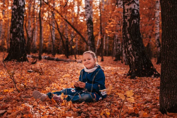 Intero concetto di infanzia felice lunghezza. sorridente bella bambina in blue jeans cappotto e jeans sedersi a terra coperto di foglie rosse cadute a fondo foresta autunno — Foto Stock