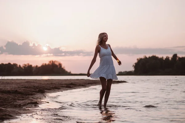 Una joven está dando vueltas en la playa. Una hermosa rubia feliz delgada en un vestido blanco de verano baila en la orilla del río sobre el fondo de la puesta de sol y las nubes —  Fotos de Stock
